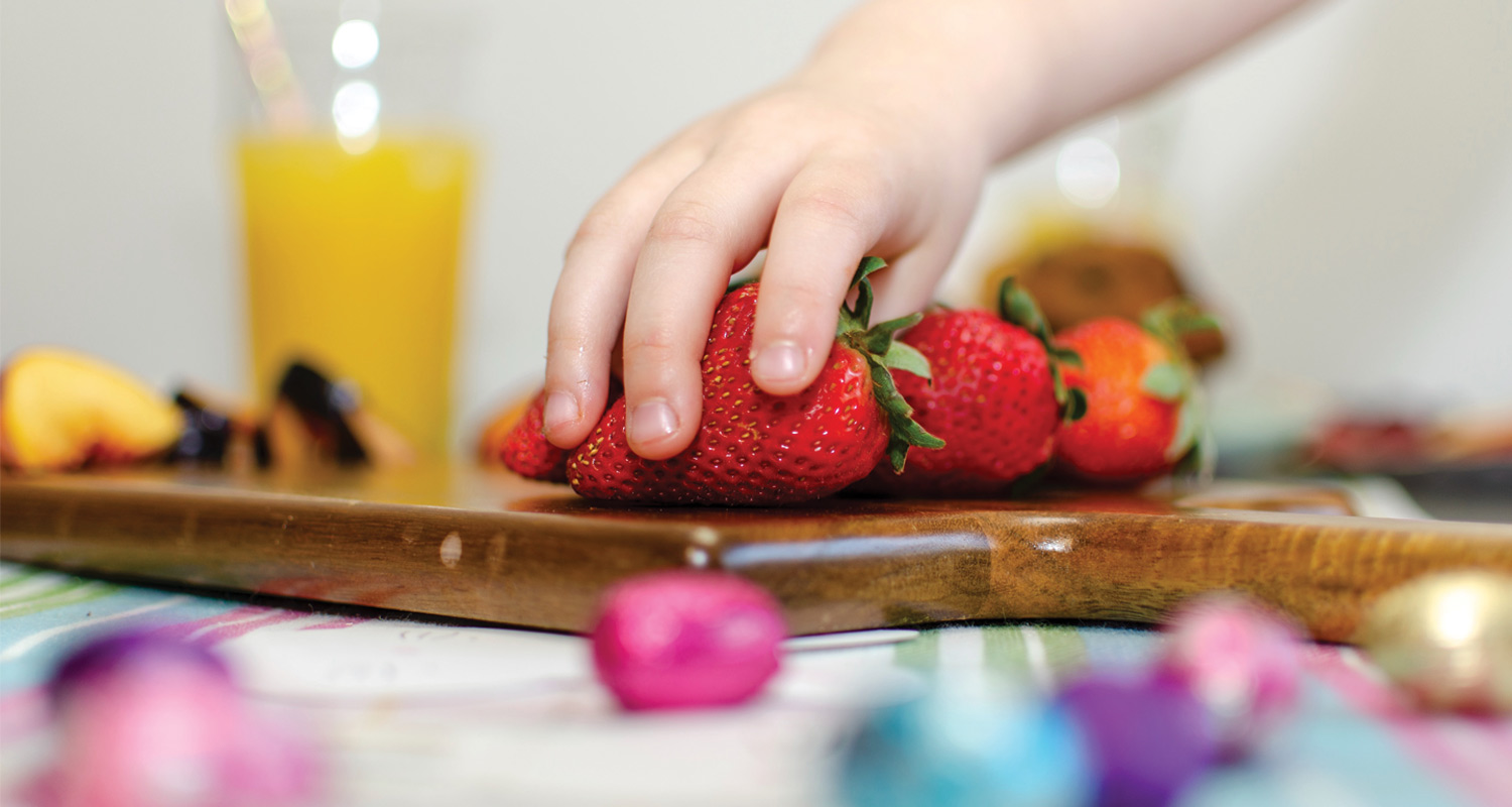a childs hand reaches for a strawberry from a pile on a cutting board