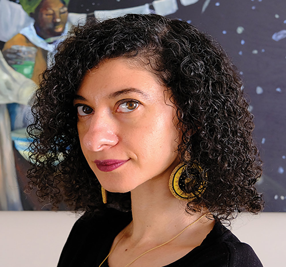 A portrait headshot picture of Chanda Prescod-Weinstein (Associate professor of physics and astronomy and core faculty member in women's studies at UNH) grinning in a dark pinkish lipstick and golden/black earrings while wearing a black shirt and a golden necklace posing in front of a painting