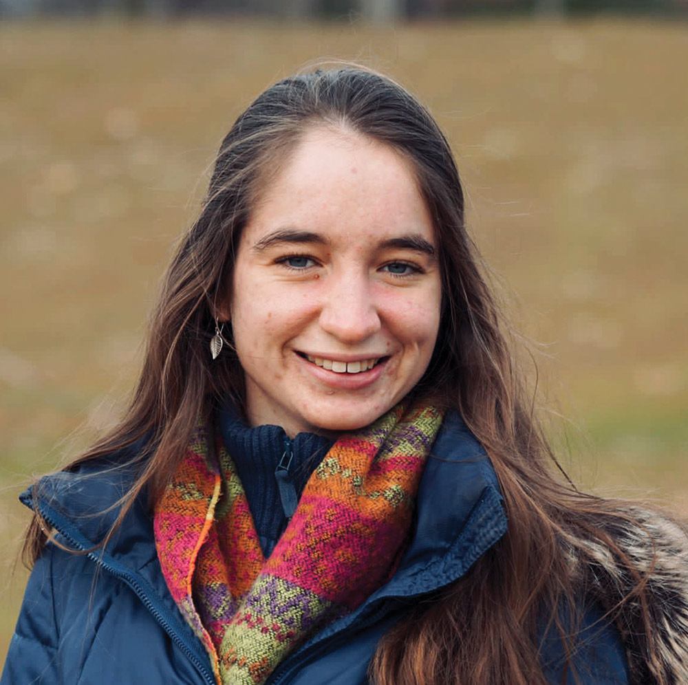 A portrait headshot picture of Grace McCulloch smiling (Master's student, natural resources) in a dark navy blue puffy jacket and a multi-colored scarf (dark pink, purple, orange, and faded red plus yellow) posing outside
