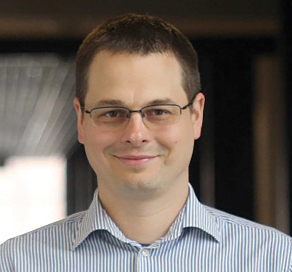 A portrait headshot picture of Marek Petrik (Assistant professor of computer science at UNH) grinning in see through prescription glasses wearing a blue and white lined pattern style button-up dress shirt with his front top collar open posing outside