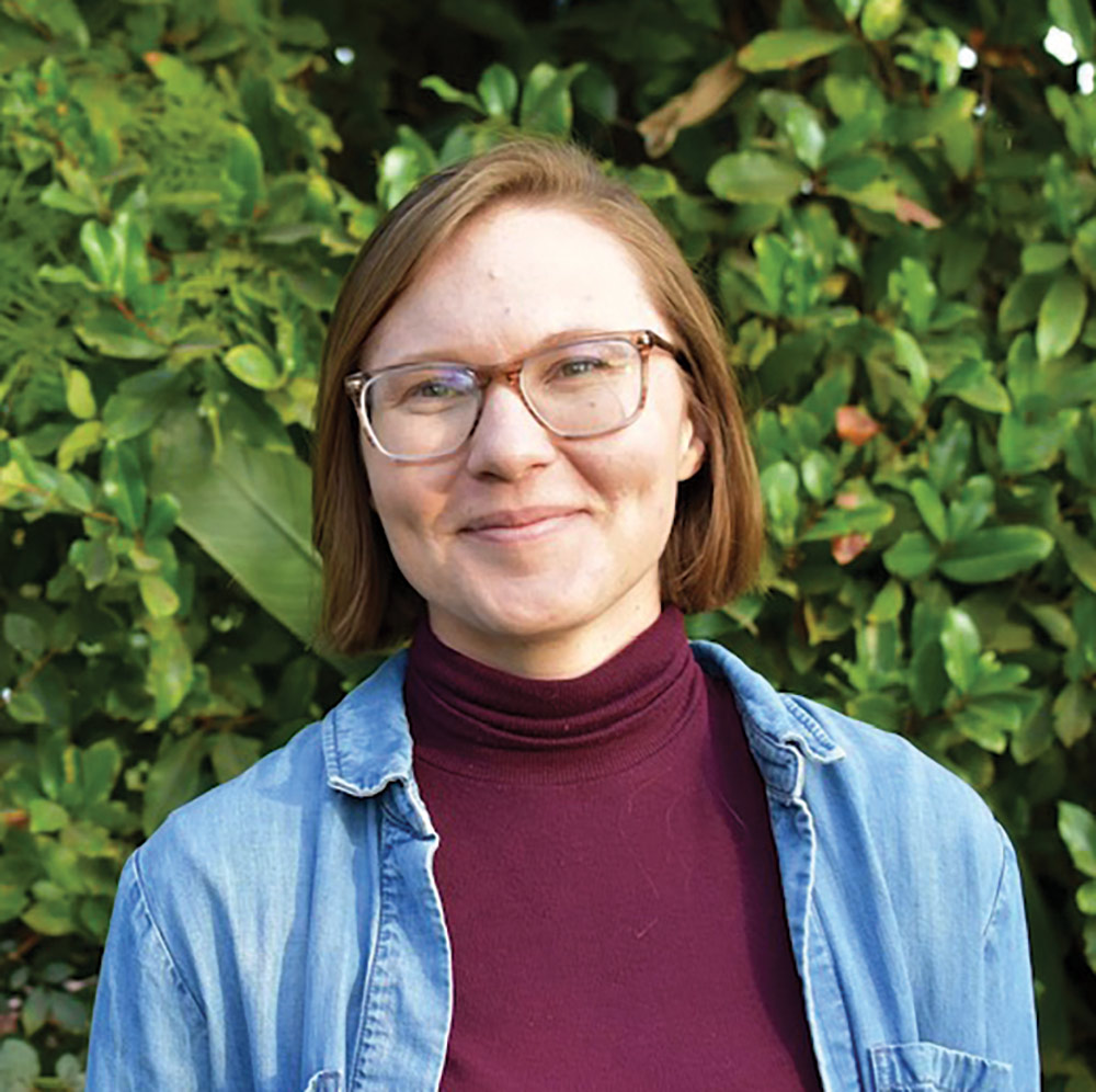 A portrait headshot picture of McKenzie Kuhn grinning (Postdoctoral researcher) in see through prescription glasses wearing a sky blue denim jacket and a burgundy turtleneck t-shirt posing outside in front of some shrubs