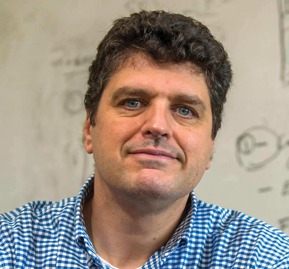 A portrait headshot picture of Nathan Schwadron (Professor of physics and astronomy at UNH) grinning in a blue and white line pattern button-up dress shirt with the front top collar open posing in front of a whiteboard
