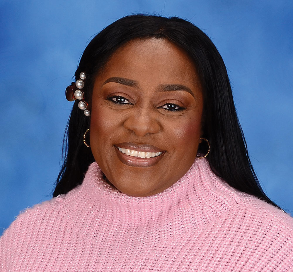 A portrait headshot picture of Ngozi Enelamah (Assistant professor of social work at UNH) smiling in a dark colored (bronze) hair clip and bronze earrings wearing a bright pink cardigan posing in front of a blue and white colored studio background