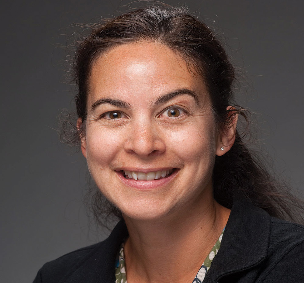 A portrait headshot picture of Semra Aytur (Professor of health management and policy at UNH) smiling in a black jacket and multi colored/pattered style dress shirt (green, blue, and white)