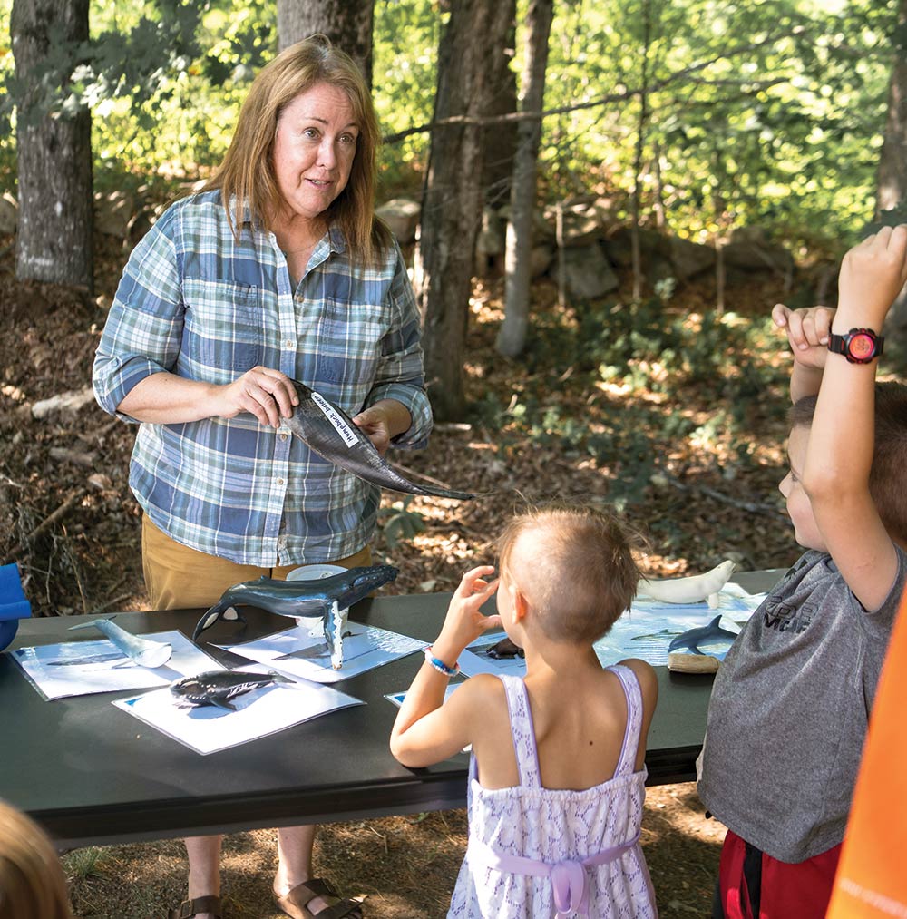 Dari Christenson (woman in center standing behind a black tabletop outside nearby some small trees in a blue colored button-up t-shirt, dark tan shorts, and dark brown sandals), a marine education program manager for NH Sea Grant, brought whale baleen samples to educate/show children and adults alike (positioned across from Dari Christenson) the importance of marine programming to public libraries