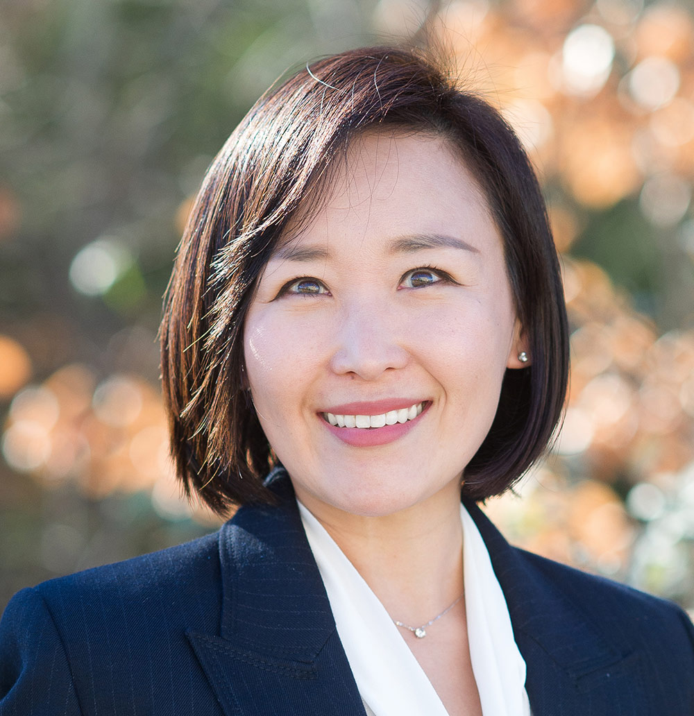 Portrait headshot close-up photo view of Hyun Ju Kim smiling in light faded pink lipstick as she has on a dark navy blue plaid business blazer coat suit with a white blouse underneath the blazer plus she is also wearing a chrome-colored necklace and earrings